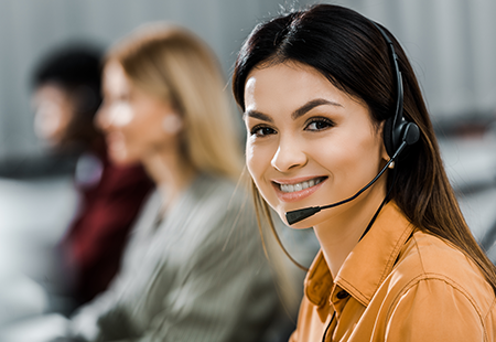 A woman with headphones smiling at the camera, with other staff members blurred in the background, indicating they are part of the contact support team.